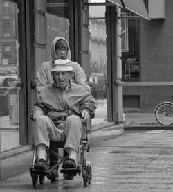 Older man in a wheelchair being pushed down the sidewalk by an older female.